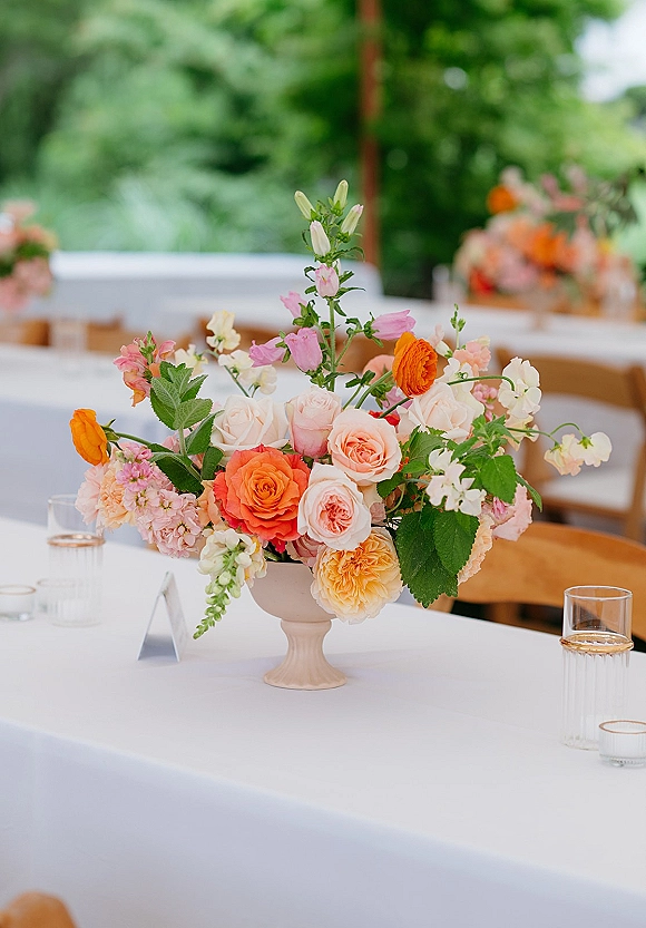 Wedding centerpiece with garden wedding centerpiece blooms of roses, ranunculus, and snapdragons in a white pedestal vase on a round table outdoors