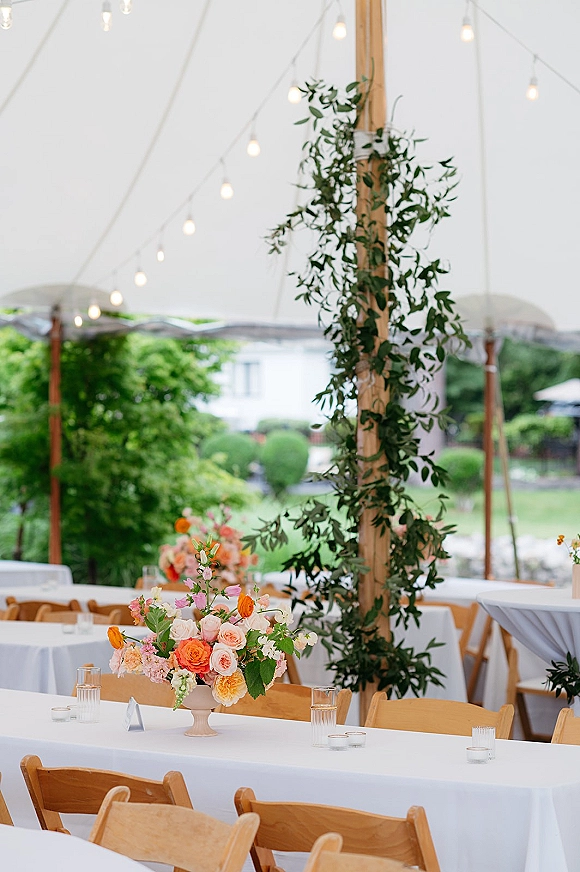 Reception tablescape under an outdoor tent reception with white linens, pedestal floral centerpiece, glass tumblers, votive candles, and string lights