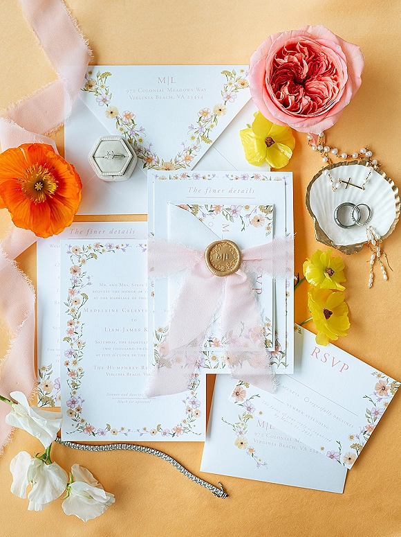 Invitation suite with floral wedding invitations, vellum wrap and wax seal, pink ribbon, rings and pearl necklace on a peach backdrop
