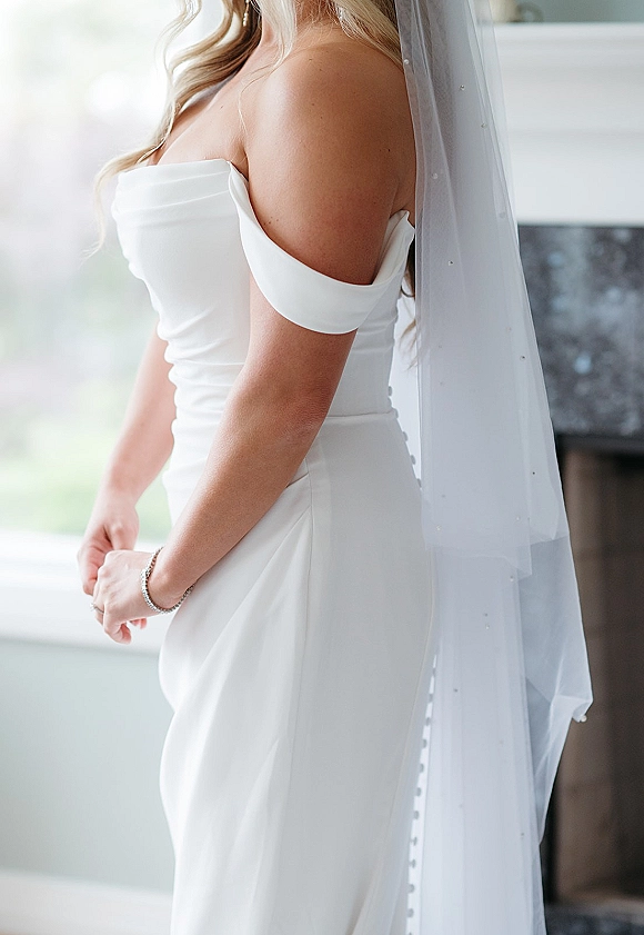 Bridal portrait of a bride in an off the shoulder wedding dress with long veil by window light near a fireplace mantel