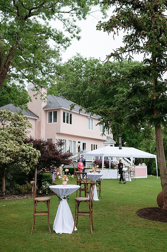 Outdoor cocktail hour with wedding cocktail hour setup featuring high top tables in white linens, pink vase florals, and a canopy bar on a lawn by a house deck