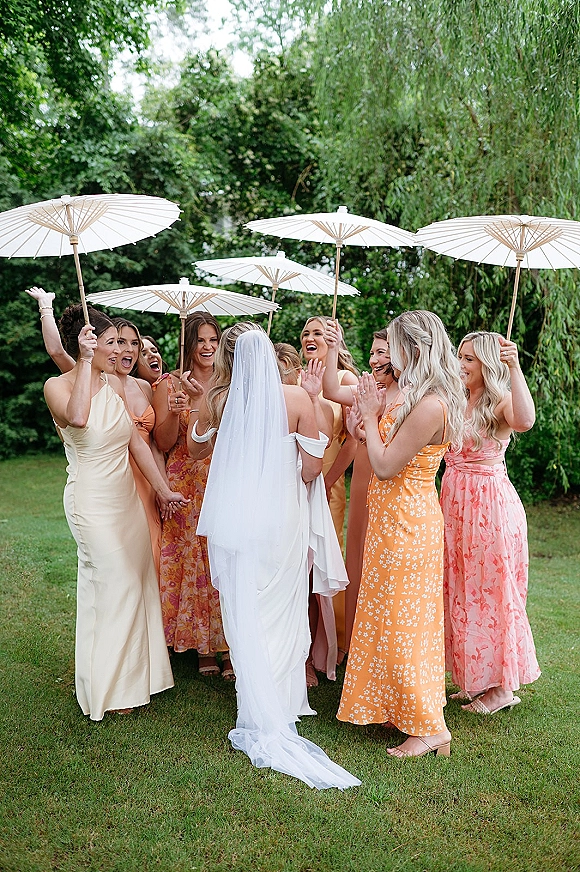 Bride with bridesmaids cheering bride as she walks on grass, cathedral veil and long train flowing in a garden, one white parasol raised