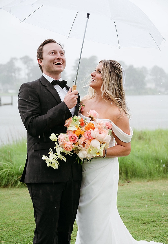 Couple portrait in rainy wedding photos, smiling under a clear umbrella by a lake, bride holding a colorful bouquet on a grassy lawn