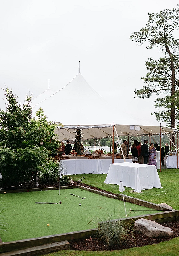 Wedding reception tent with sailcloth wedding tent string lights, cocktail tables, bar, and mini golf putting green on a lakeside lawn