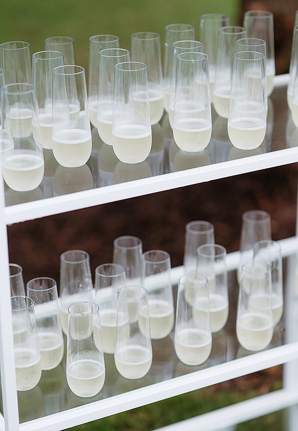 Champagne glass display with rows of pre-poured sparkling wine flutes on clear acrylic shelving and a white frame on an outdoor lawn