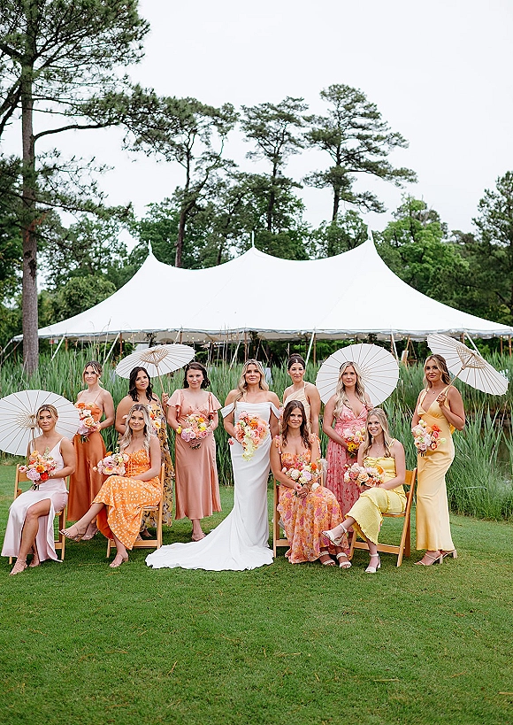 Bride with bridesmaids in colorful dresses holding bouquets, some with white parasols, seated on wooden chairs under a sailcloth tent
