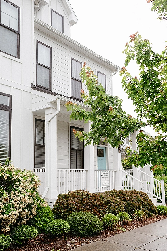 Wedding venue exterior of a white house wedding venue with porch columns and a turquoise front door, lined by flowering shrubs under an overcast sky