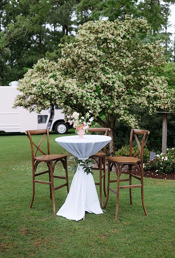Cocktail table setup with white tablecloth, wooden bar stools, and small pink-vase floral centerpiece on a garden lawn under flowering trees