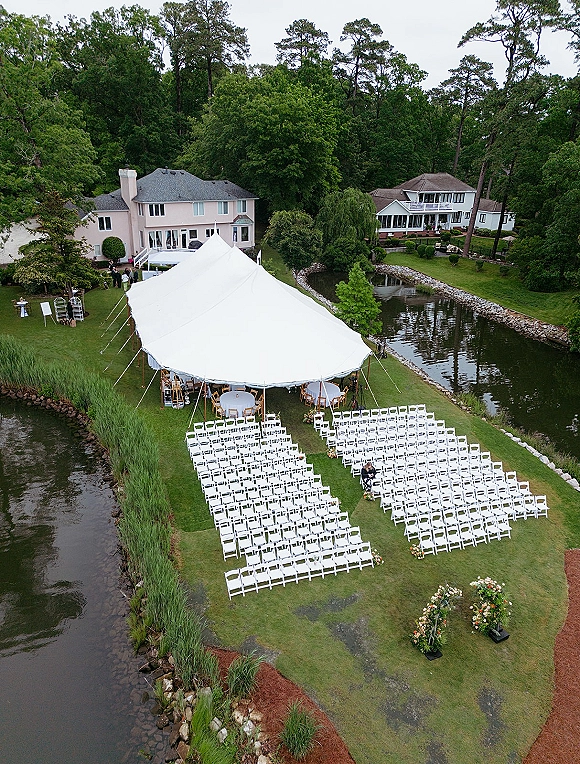 Outdoor ceremony setup with a backyard wedding ceremony under a white tent, folding chairs facing a floral arch by the river