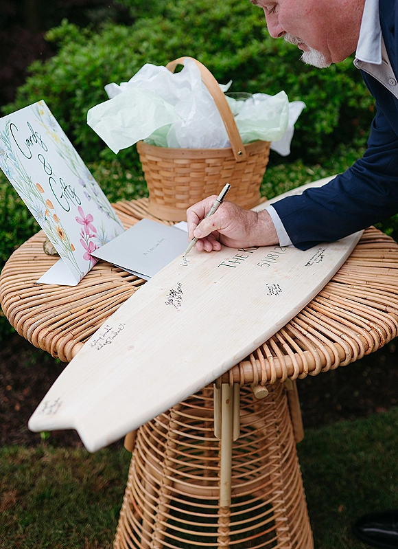 Wedding guest book surfboard guest book on a wicker table with a black pen, greeting card, and basket, set on a garden lawn