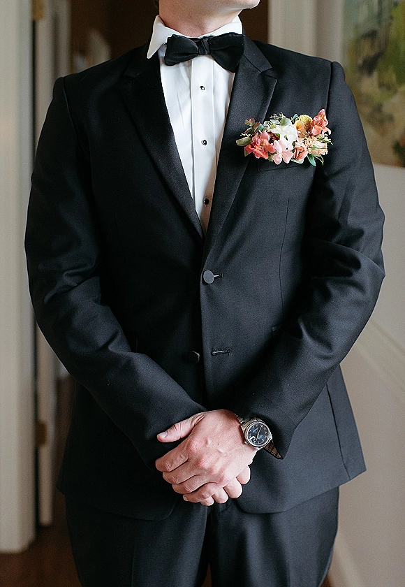 Groom portrait in a black tuxedo groom look, hands clasped with watch, bow tie and boutonniere in an indoor hallway with wall art