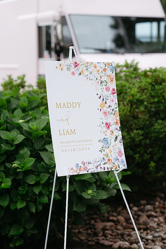 Wedding welcome sign with a floral border and gold lettering on a white easel, set on gravel near green shrubs and a white vehicle