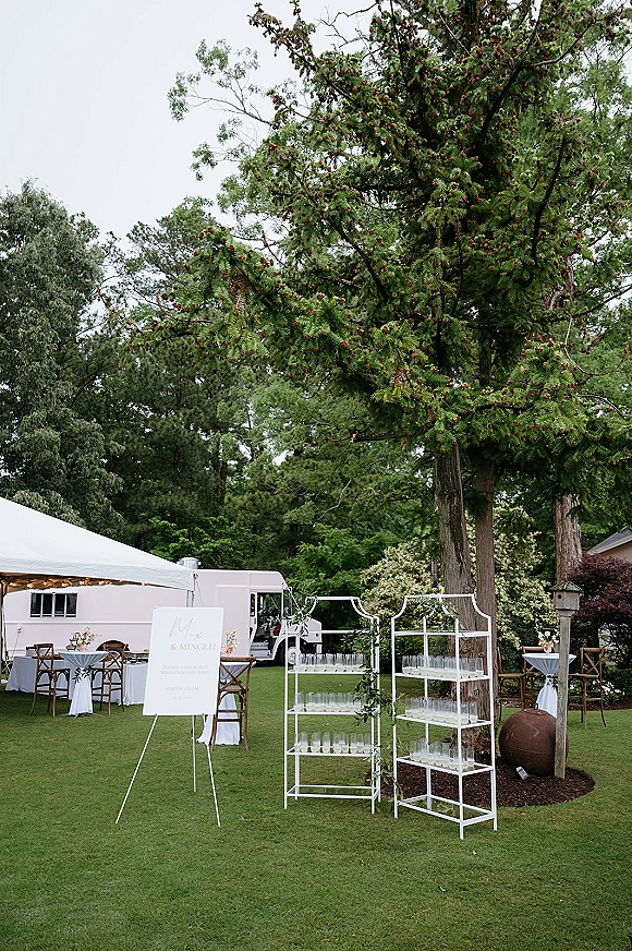 Outdoor wedding cocktail hour setup with linen cocktail tables, wooden chairs, welcome sign, and trailer bar under a white tent with string lights