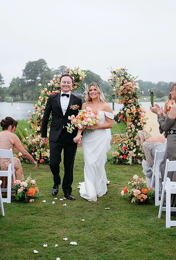 Wedding recessional as bride and groom walk the aisle, bouquet in hand, passing rose petals and guests with a lakeside backdrop under overcast sky