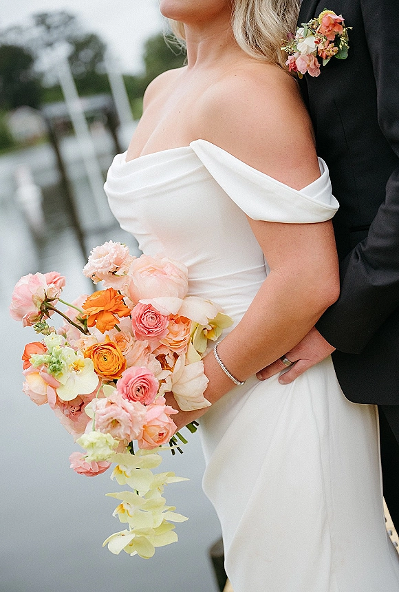 Couple portrait of bride and groom embrace on a waterfront dock, her off the shoulder dress and bouquet of roses and orchids under overcast sky