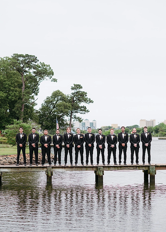 Groomsmen portrait in matching black tuxedos with bow ties and boutonnières lined up on a wooden dock by a lake under cloudy skies