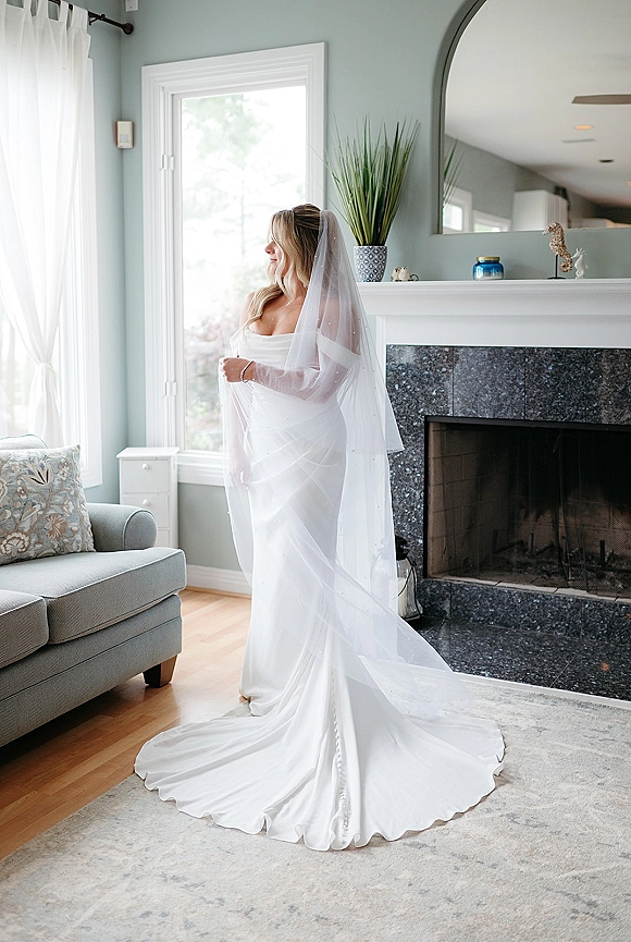 Bridal portrait of a bride by window in a strapless wedding dress with long veil and gown train, standing in a cozy living room near a fireplace
