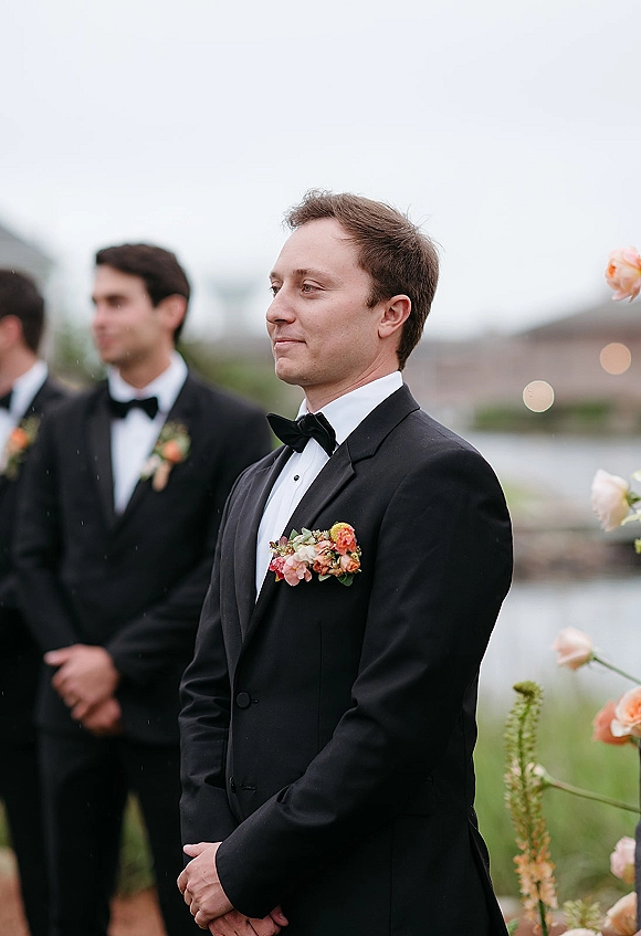Groom portrait in a black tuxedo with bow tie and peach boutonniere, standing by a floral arch at an overcast waterfront ceremony