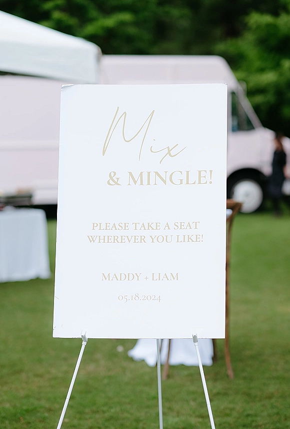 Wedding welcome sign with gold lettering on a white board easel, set on a grassy lawn near trees, an event tent, and pink trailer