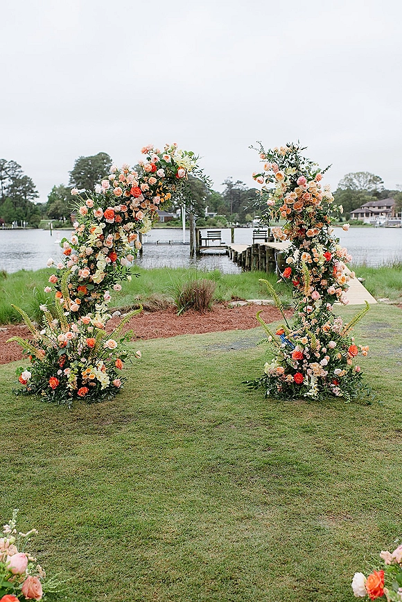 Wedding ceremony arch with floral ceremony arch pillars of roses and greenery, set on a dockside lawn by the waterfront under clouds
