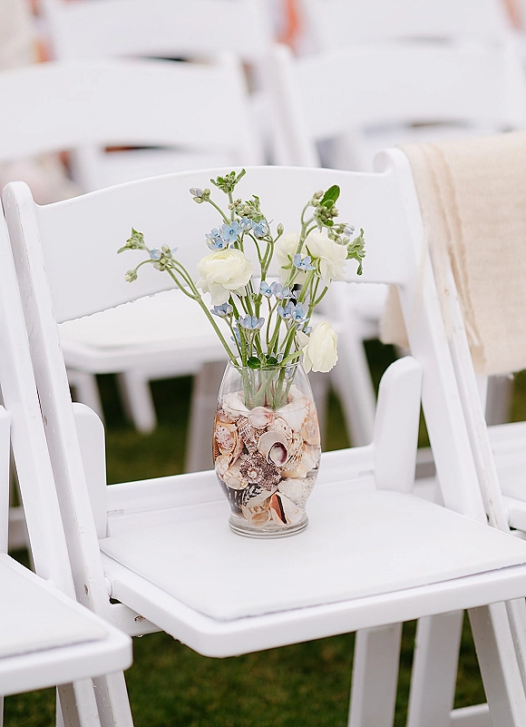 Ceremony chair decor with a glass vase of seashells and blue-white flowers on a burlap-sashed white folding chair in lawn rows