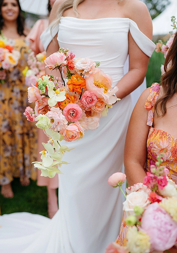 Bridal bouquet in coral and peach bouquet tones with peonies, ranunculus and orchid cascade, held against an off-shoulder gown outdoors