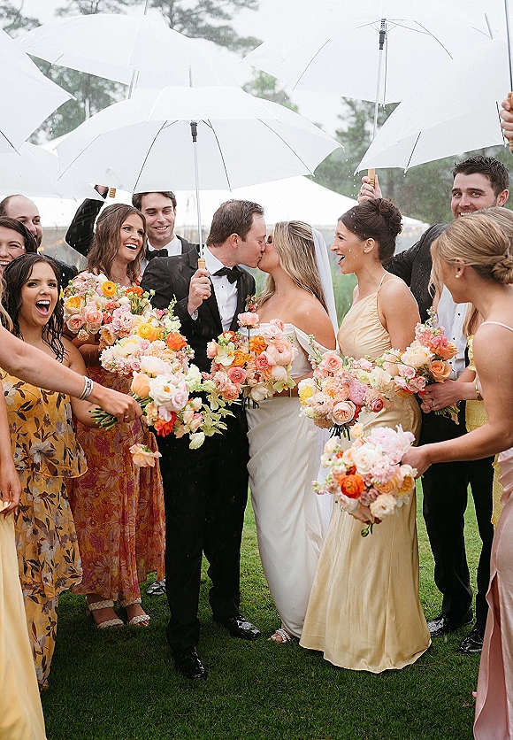 Wedding kiss in the rain as bride and groom under umbrella, bridal party holds white umbrellas and bouquets on a grassy lawn with trees