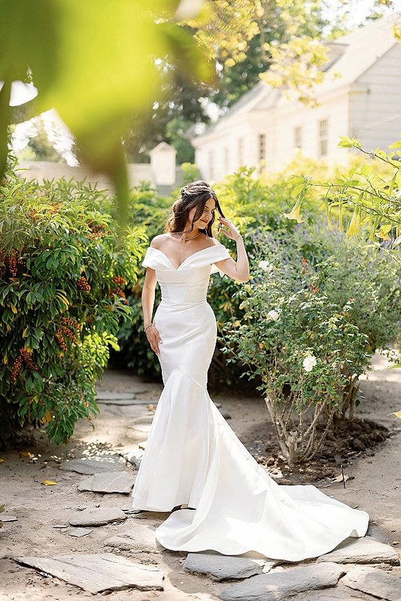 Bridal portrait of a bride in an off the shoulder wedding dress with long train, looking down on a stone path amid garden greenery