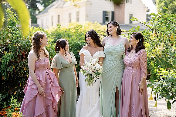 Bride with bridesmaids posing in pastel dresses, holding a classic bridal bouquet in front of garden greenery and a house exterior