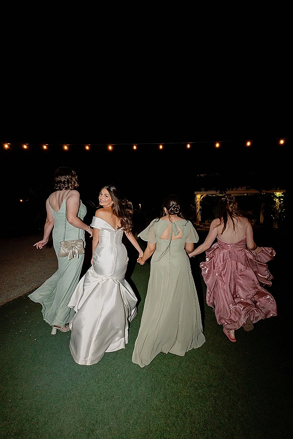 Bridal party photo of the bride with bridesmaids walking away on a lawn at night under string lights, bride looking back in satin gown