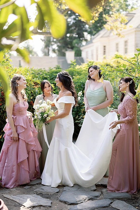 Bride with bridesmaids helping bride gather her long dress train on a sunlit garden stone path, holding a white bouquet near a house exterior