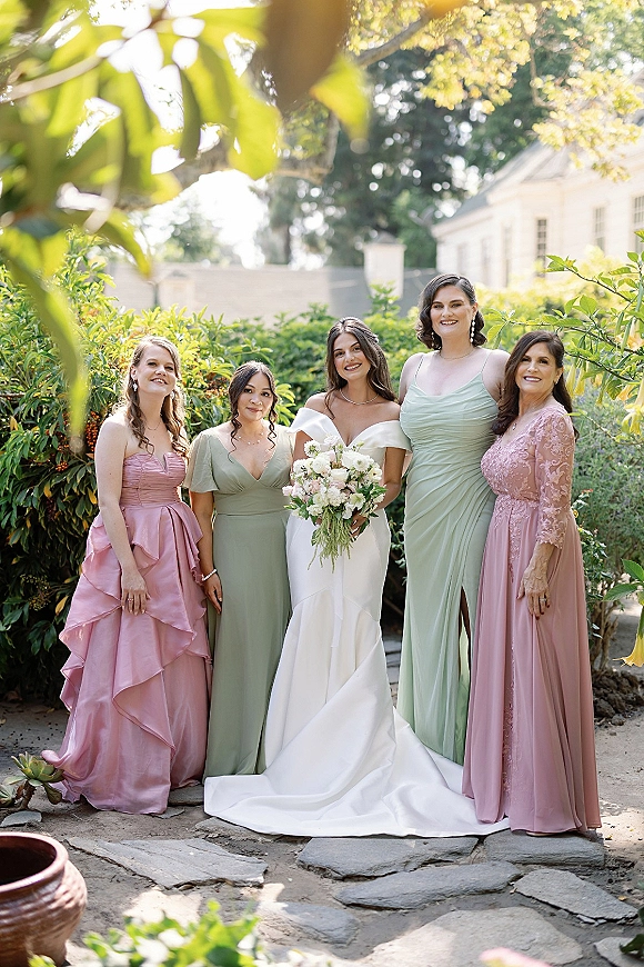 Bridesmaid group photo of bride with bridesmaids in sage and dusty rose dresses, holding white rose bouquet on a sunlit garden path