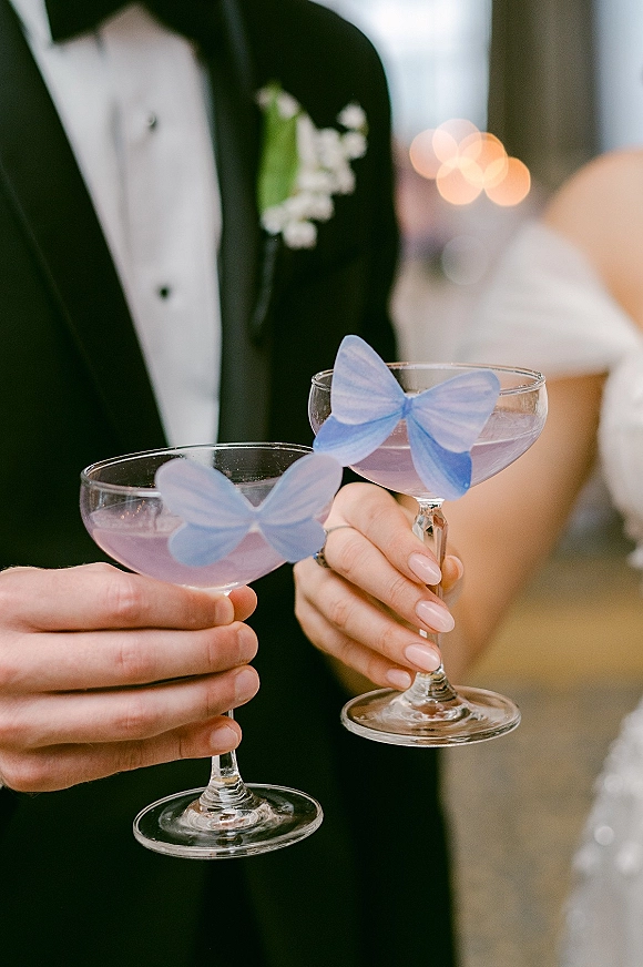 Wedding cocktail toast as bride and groom clink coupe glass cocktails with purple drinks, butterfly toppers, rings and bokeh reception lights