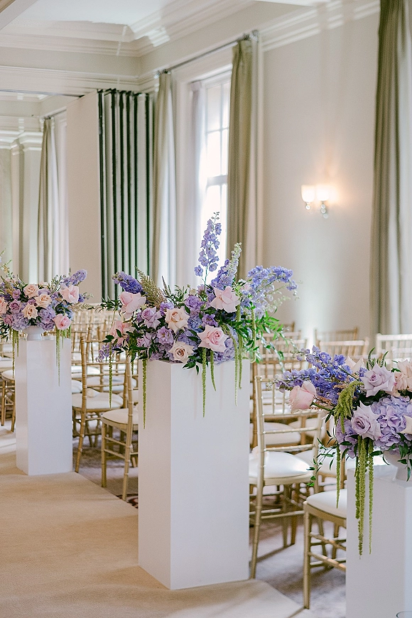 Ceremony aisle decor with wedding aisle flower pedestals on white stands, lavender and blush roses, gold chiavari chairs in a bright ballroom