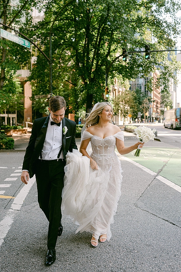 Couple portrait of bride and groom walking across a city street crosswalk, bride holding a bouquet and dress train beside him