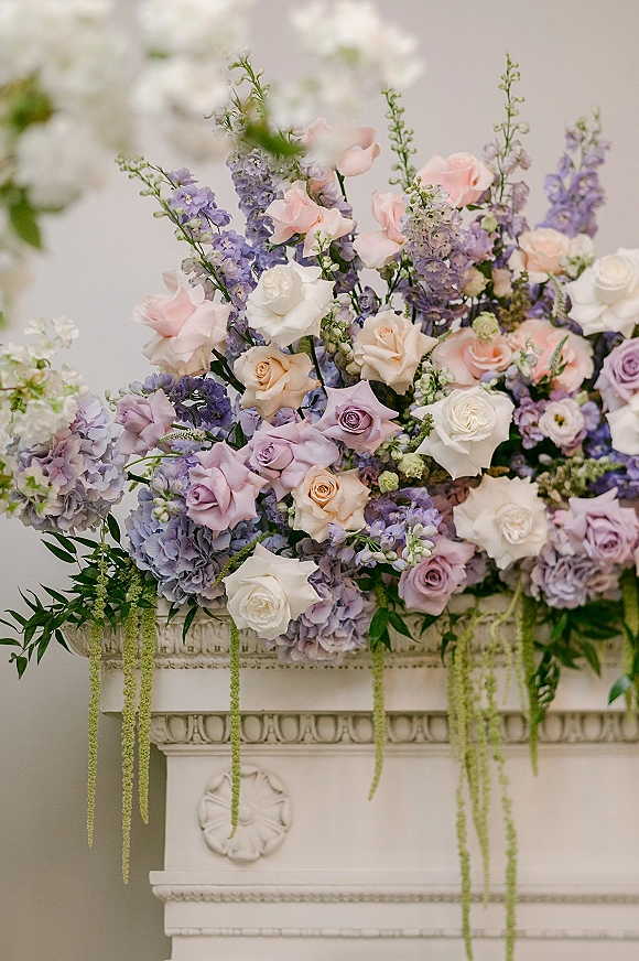 Wedding floral arrangement with roses and hydrangeas cascading across a white mantel, with hanging amaranthus and greenery against a neutral wall