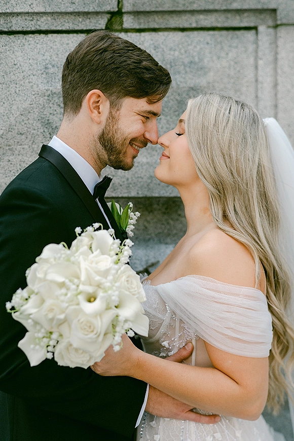 Wedding couple portrait with bride and groom close up, forehead touching beside a stone wall, bride holding white calla lily bouquet and veil