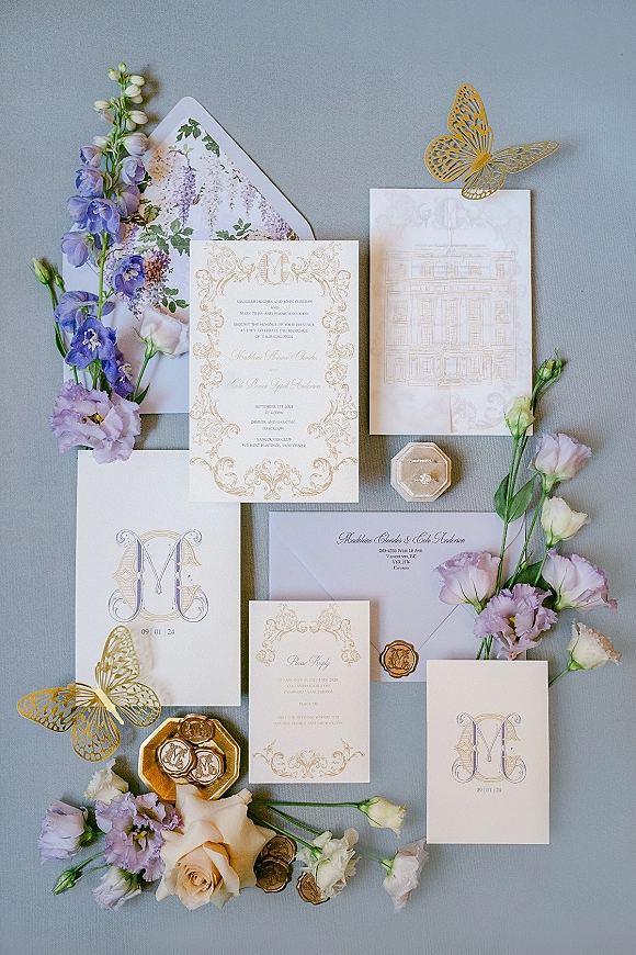 Wedding invitation suite with gold foil wedding invitations, wax seals, butterfly cutouts, ring box, and pastel blooms on gray backdrop