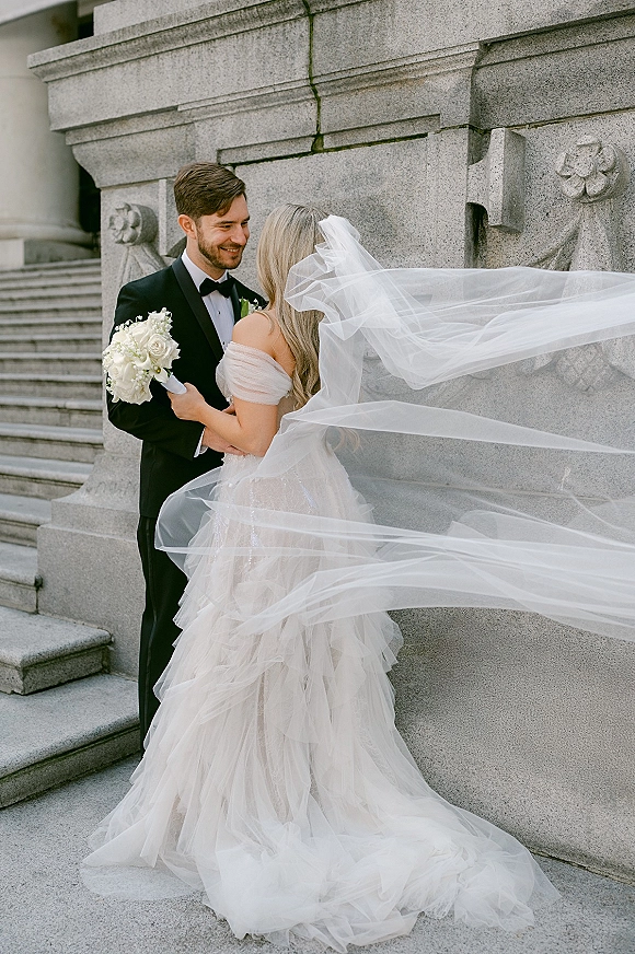 Couple portrait of bride and groom embrace on stone steps, her long cathedral veil blowing as she holds a white rose bouquet