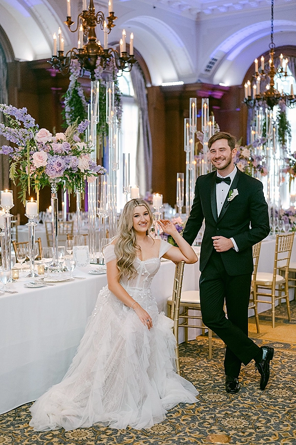 Couple portrait of bride seated in an off-the-shoulder ball gown and groom in a tuxedo beside candlelit reception tables in a chandelier ballroom