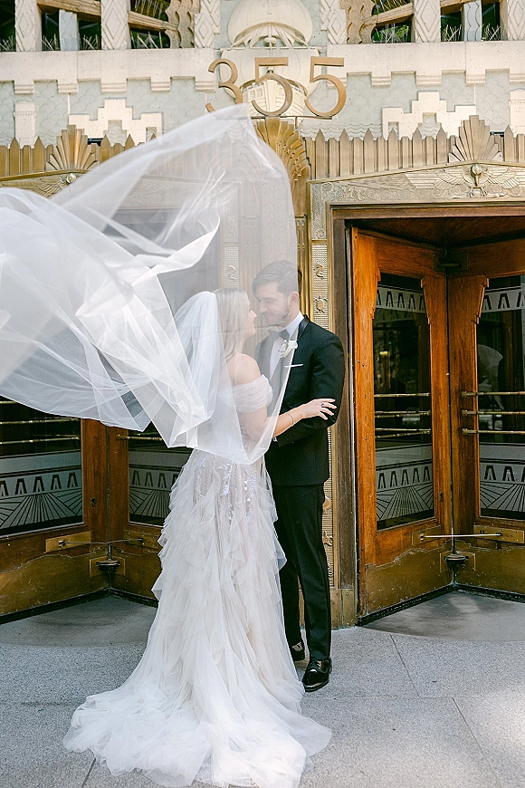Couple portrait of bride and groom portrait embracing under a flowing wedding veil at an art deco entrance with revolving door and marquee numbers