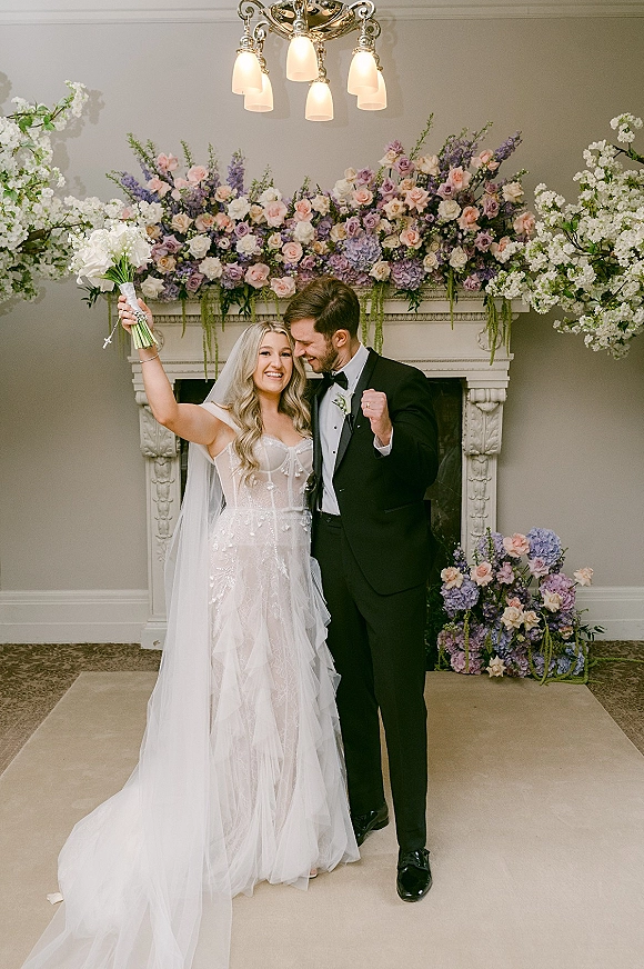 Wedding couple portrait of bride holding bouquet and groom in black tuxedo embracing by a floral fireplace mantel with chandelier overhead