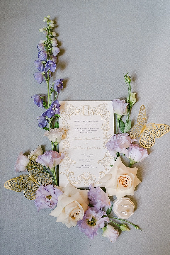 Invitation suite wedding invitation flat lay with purple delphinium, ivory roses, gold butterfly cutouts and ring box on gray fabric backdrop