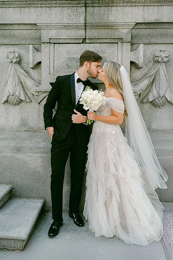 Wedding kiss portrait of bride and groom kissing, bride holding a white rose bouquet, veil flowing by carved stone wall and steps