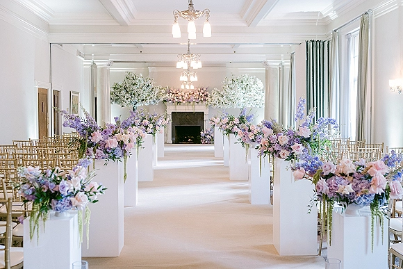Indoor ceremony setup with wedding aisle floral pillars on white pedestal plinths, gold chiavari chairs, and chandeliers framing a fireplace backdrop
