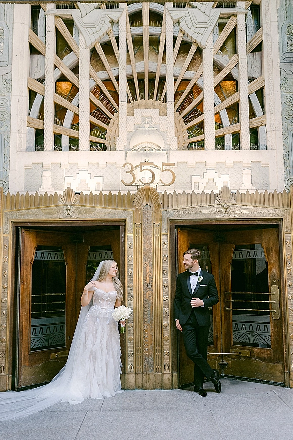 Couple portrait of bride and groom looking at each other, bride in veil holding white bouquet, by an art deco entrance with ornate doors