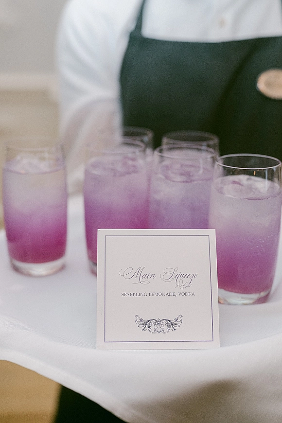 Signature cocktail sign with calligraphy drink sign beside clear highball glasses of purple iced cocktails on a white tablecloth, server blurred behind