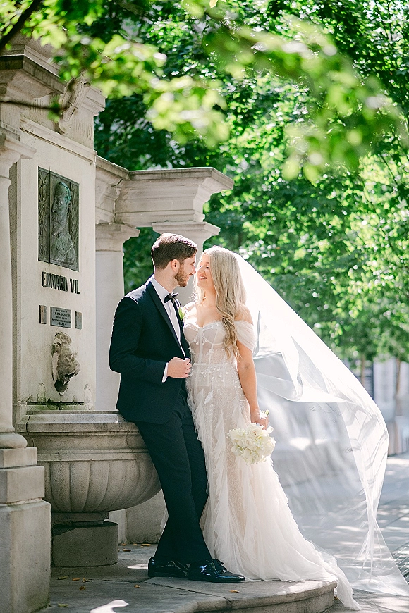 Couple portrait of bride and groom portrait with her veil blowing, holding a white bouquet beside a stone fountain on a tree-lined sidewalk