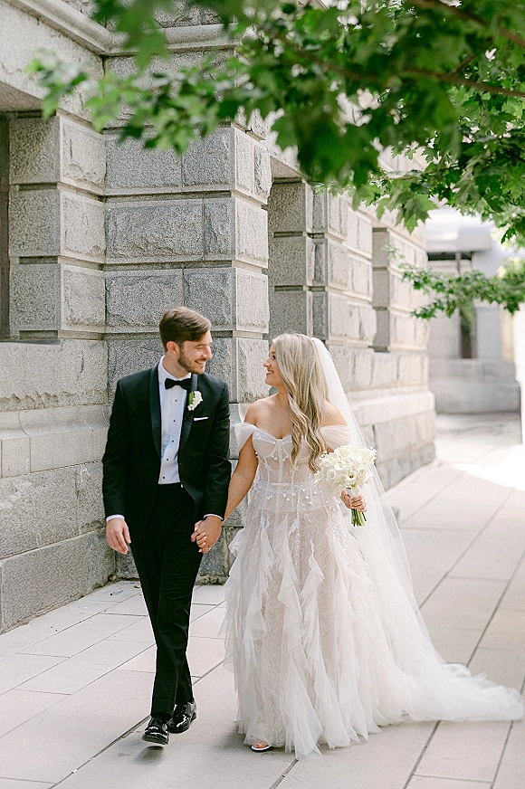 Wedding couple portrait of bride and groom walking hand in hand, bride looking at groom, veil and bouquet by a stone wall sidewalk