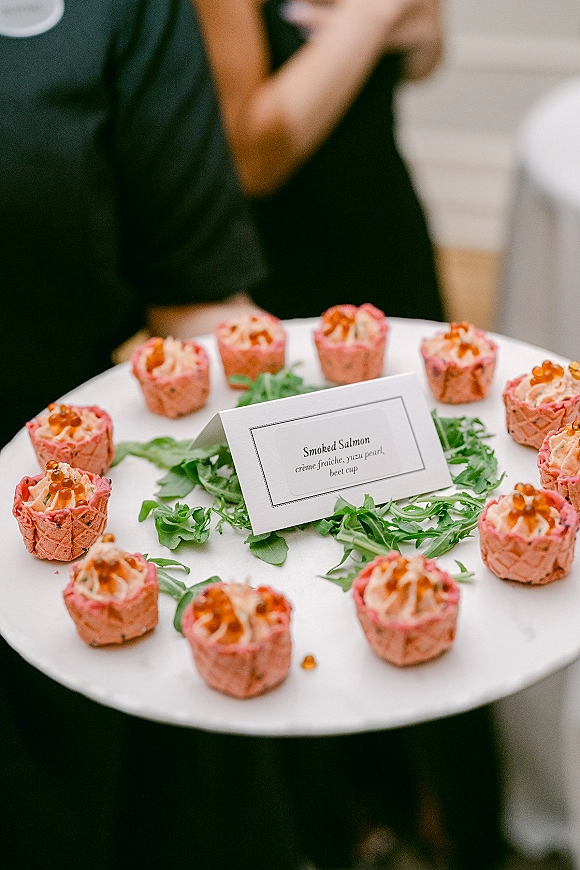 Cocktail hour appetizers on a passed hors d'oeuvres tray with smoked salmon bites, roe and arugula garnish beside a menu card